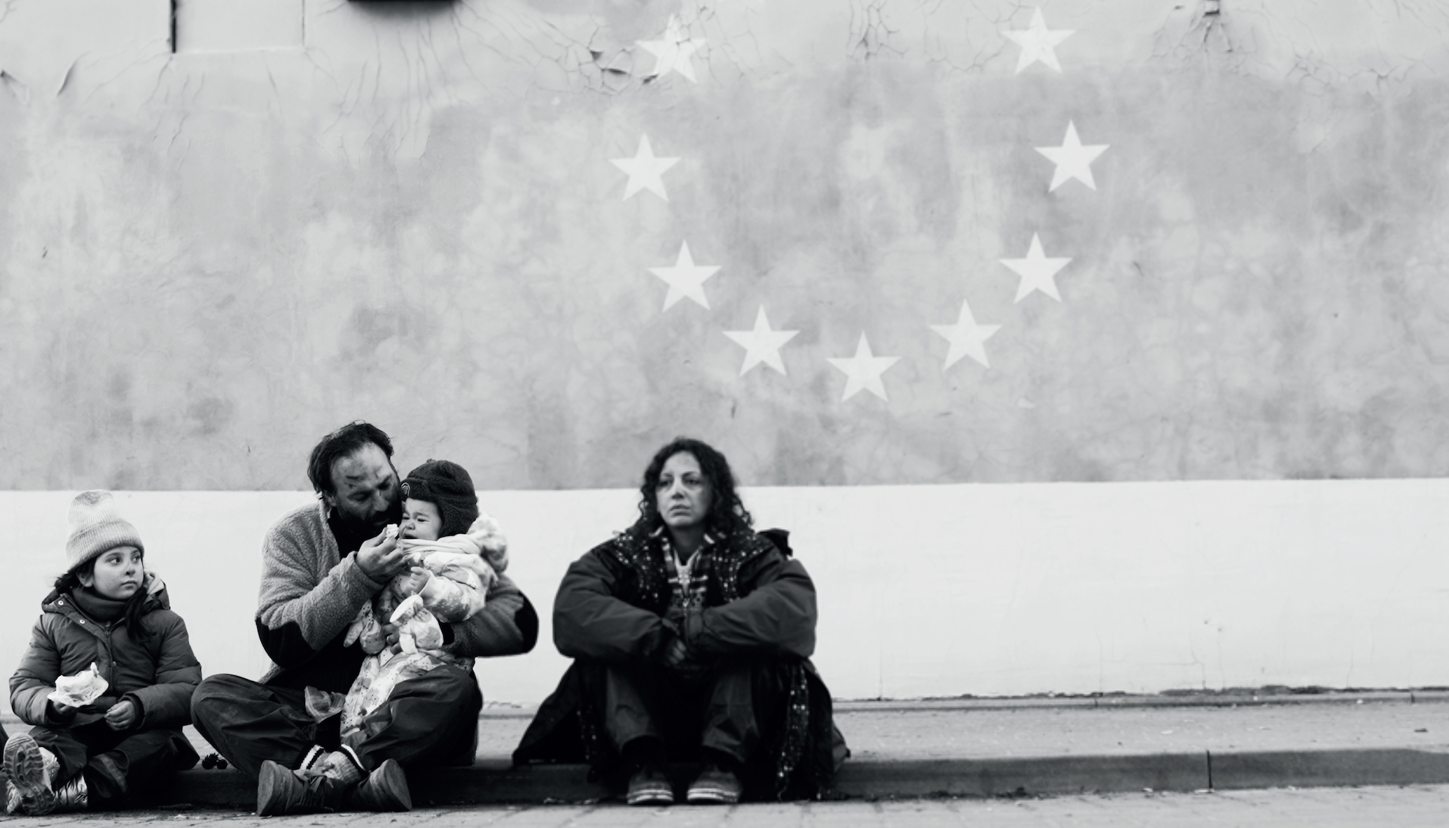 A family of four sits on the ground wearing winter coats in front of a wall with a circle of stars on it.