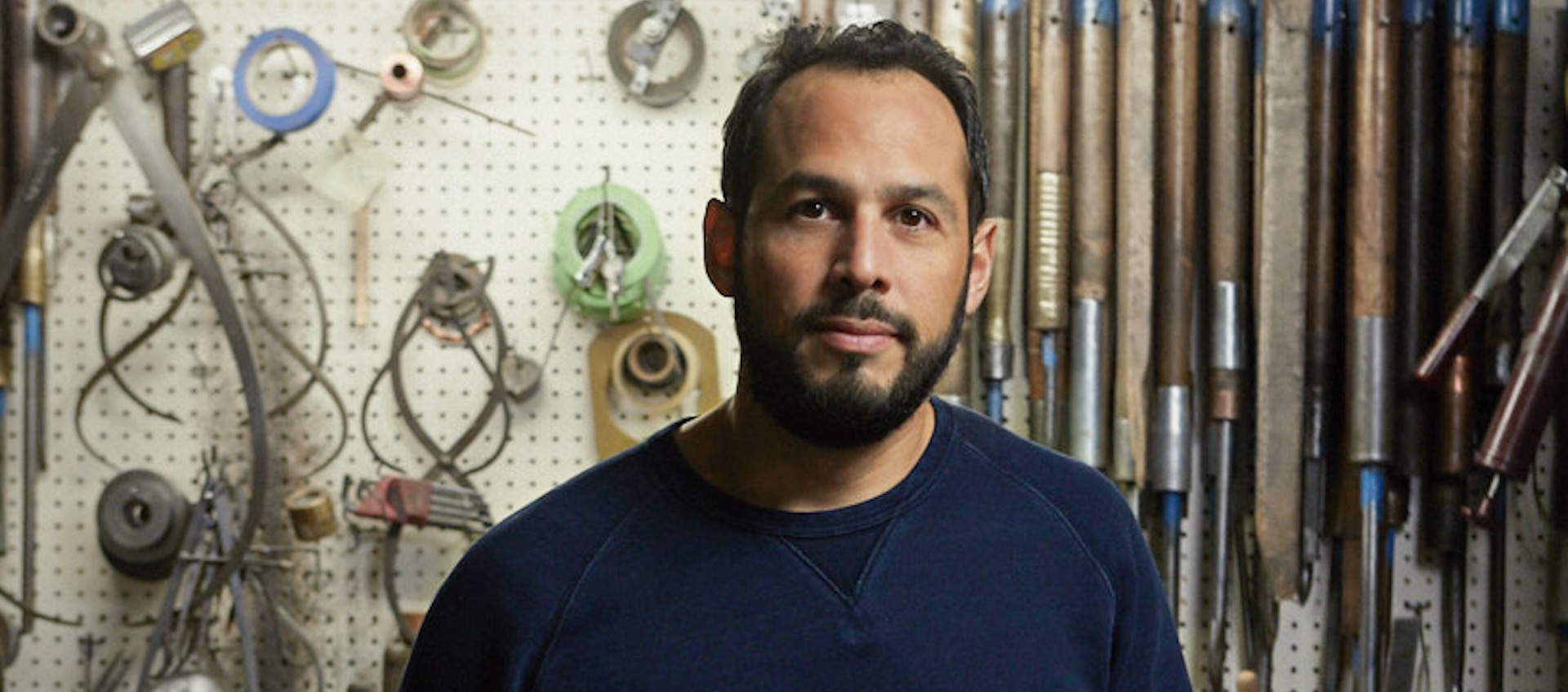 A man of Central American descent stands in his art studio and looks at the camera.
