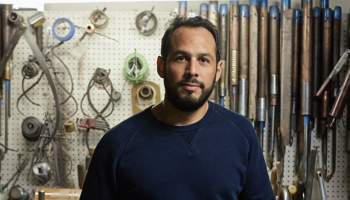 A man of Central American descent stands in his art studio and looks at the camera.