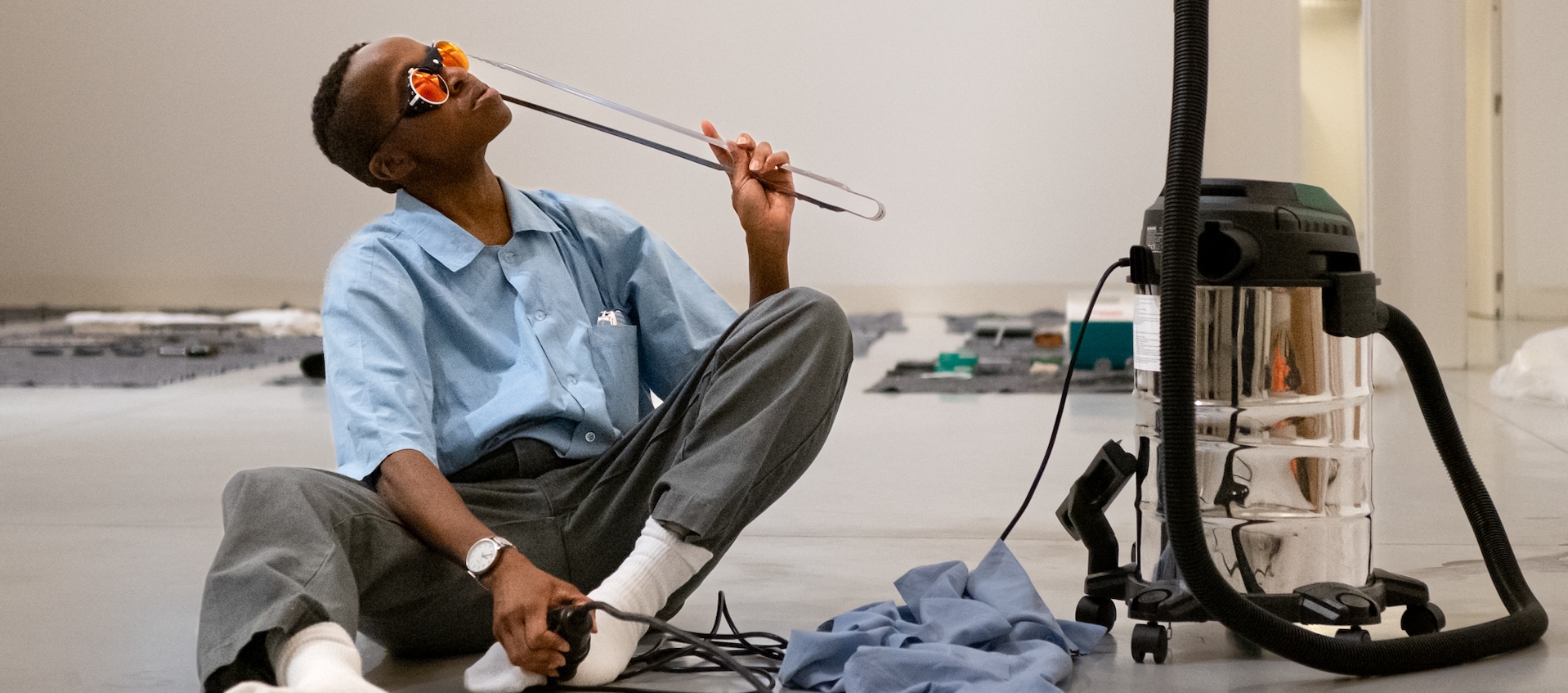 A person with dark skin, close-cropped hair and sunglasses sits in a white gallery space, holding tongs close to their face. A large canister vacuum is next to them.