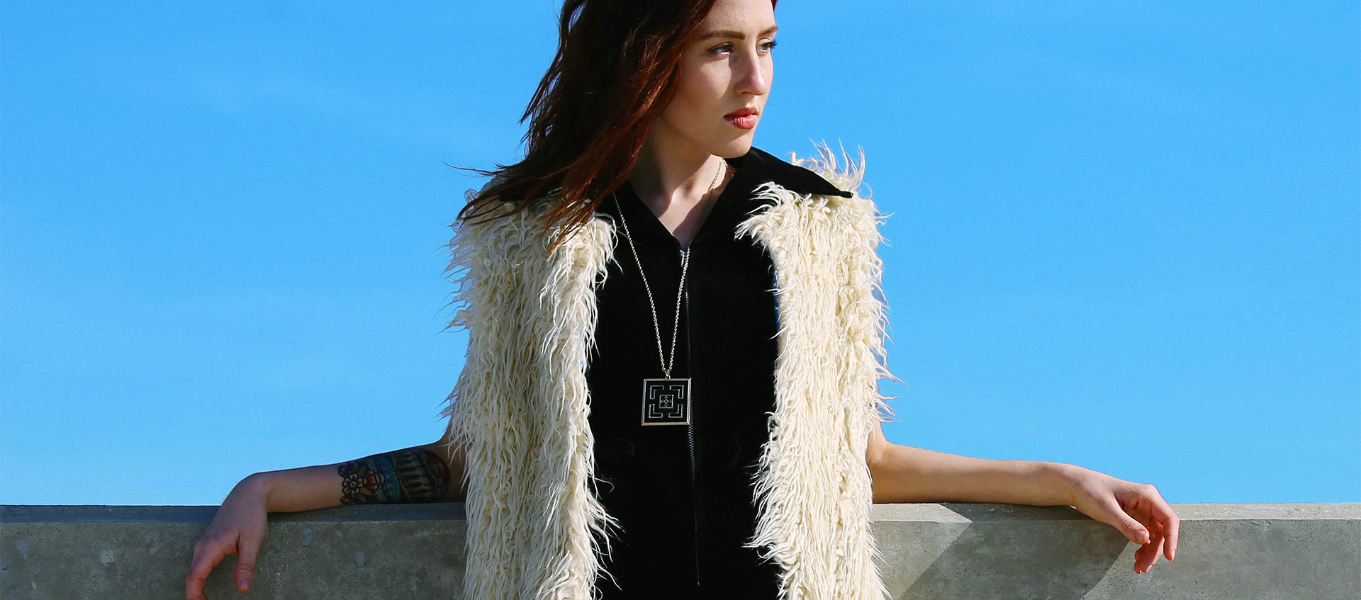 a woman in black and fur vest leaning on concrete ledge against blue sky