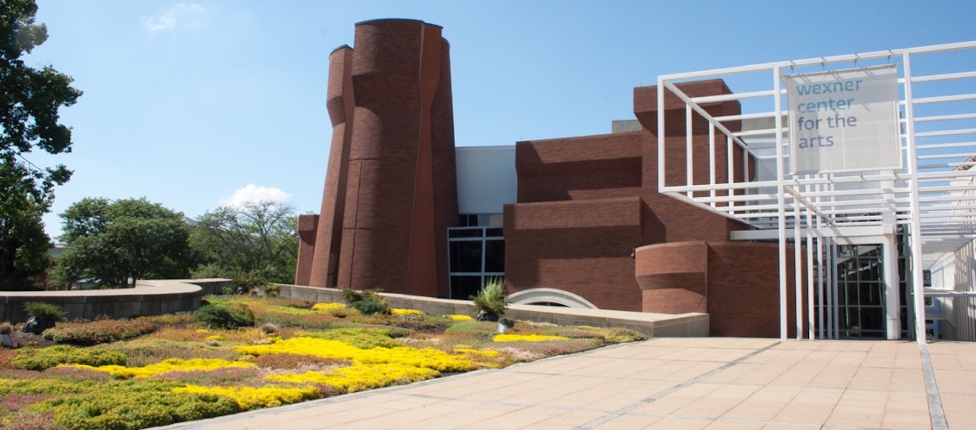 an exterior photograph of the south-facing side of the Wexner Center for the Arts at the Ohio State University, showing the deconstructed brick turrets, the external white metal grid, and an outdoor garden installation by Paula Hayes