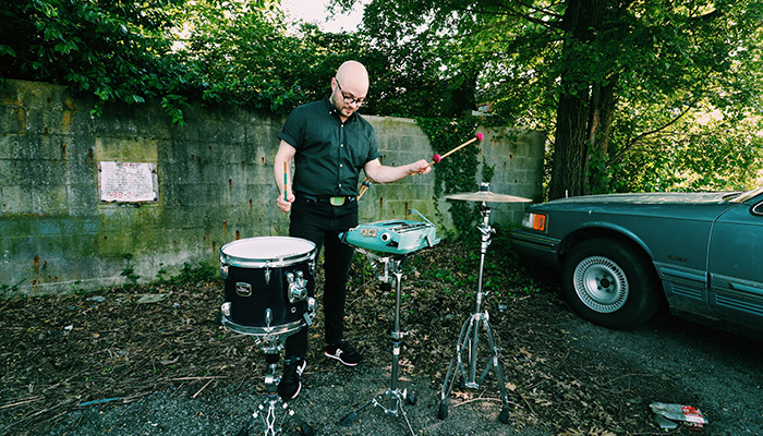 Noah Demland uses mallets to play a tom, hi-hat cymbal, and typewriter outdoors in front of cinder block and vines