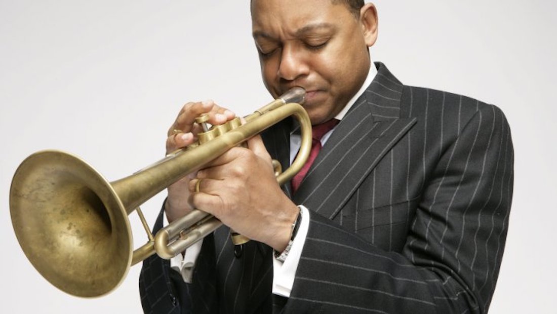 Jazz artist Wynton Marsalis seen in three-quarter profile from the chest up, playing the trumpet, against a solid light gray background