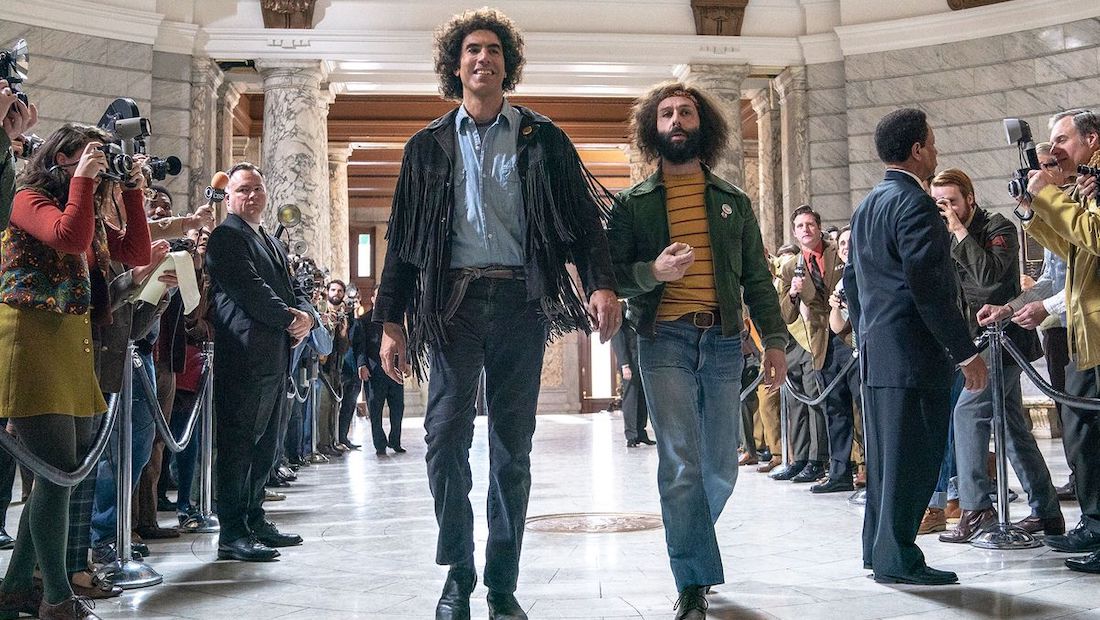 Sacha Baron Cohen as Abbie Hoffman and Jeremy Strong as Jerry Rubin walk down through a courthouse lobby as reporters behind stanchions on either side of them take pictures in a scene from the film The Trial of the Chicago 7