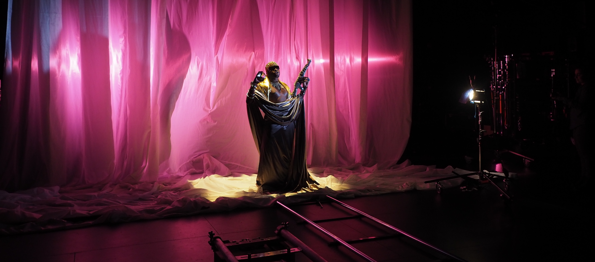 Artist Jaamil Olawale Kosoko stands against a curtain under pink lighting, holding a foil-covered toy machine gun, as he shoots a short film for his American Chameleon project