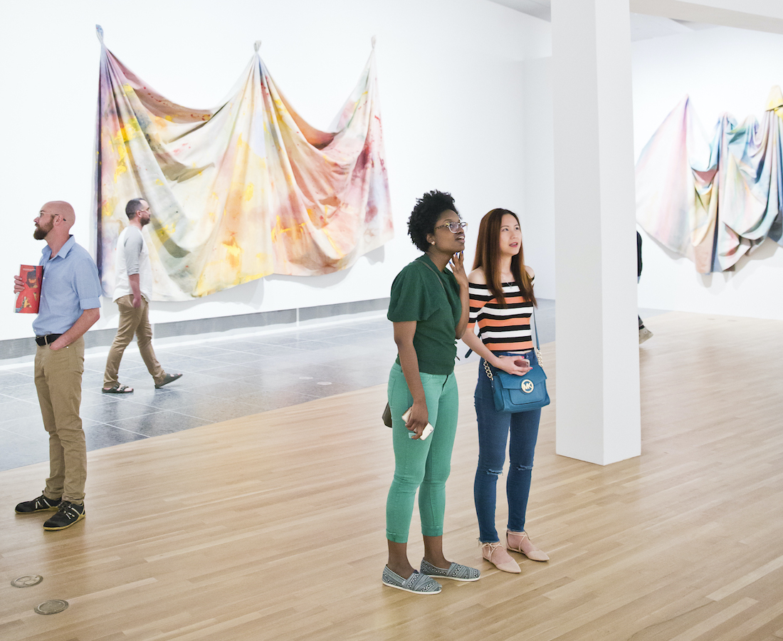 Two young women look at an unseen artwork with two draped canvases by Sam Gilliam on walls in the background during the opening of the Wexner Center exhibition Inherent Structure