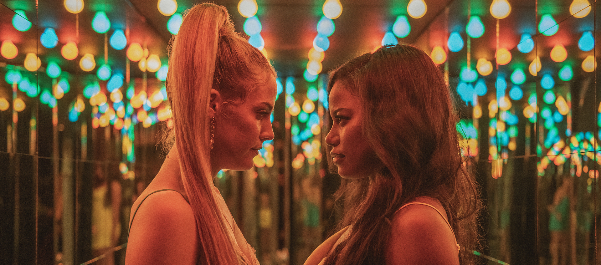 Two women stand in front of a mirrored surface and beneath rows of multicolored lights. The woman on the left has long blond hair in a ponytail and the woman on the right wears a pink top. They are both staring each other down.