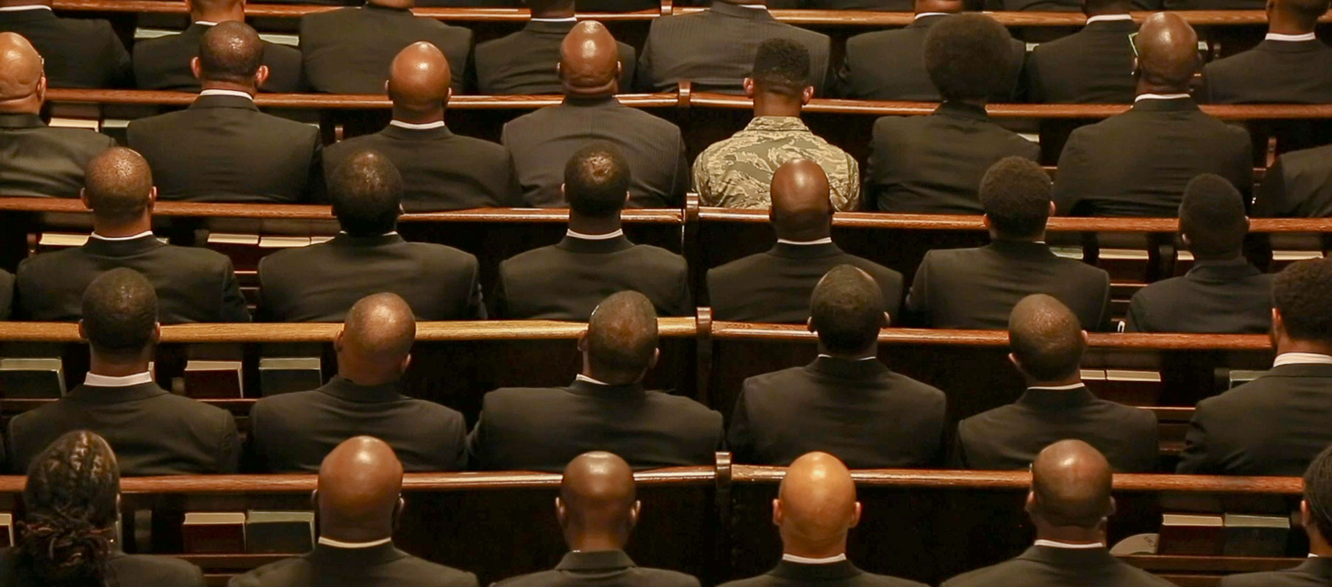 several people in dark suits, one in military fatigues, seated in pews