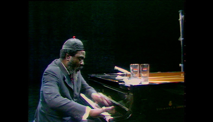 Thelonious Monk, viewed from the side, playing piano. He wears a dark suit and a black cap.