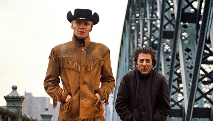 Two men are walking along a bridge. One wears a black cowboy hat and a tan buckskin jacket, the other has his hair slicked back and is wearing a dark overcoat.
