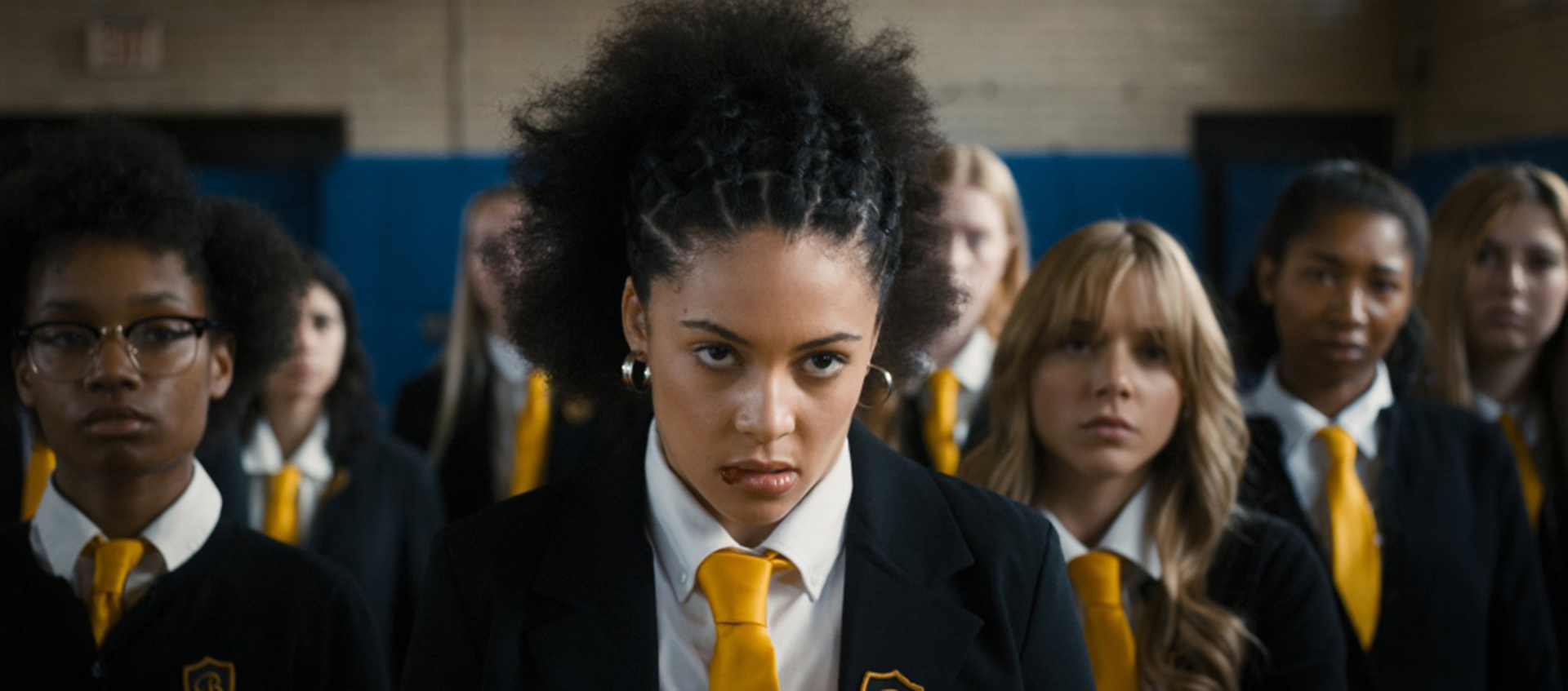 Close-up of a scowling young woman with dark, curly hair. She is surrounded by other young women in school uniforms with blue blazers and gold ties.