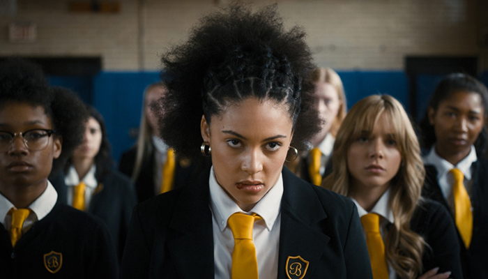 Close-up of a scowling young woman with dark, curly hair. She is surrounded by other young women in school uniforms with blue blazers and gold ties.
