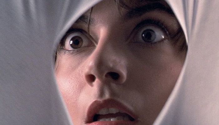 A close-up of a woman who looks startled peering through a hole in a sheet of white fabric.