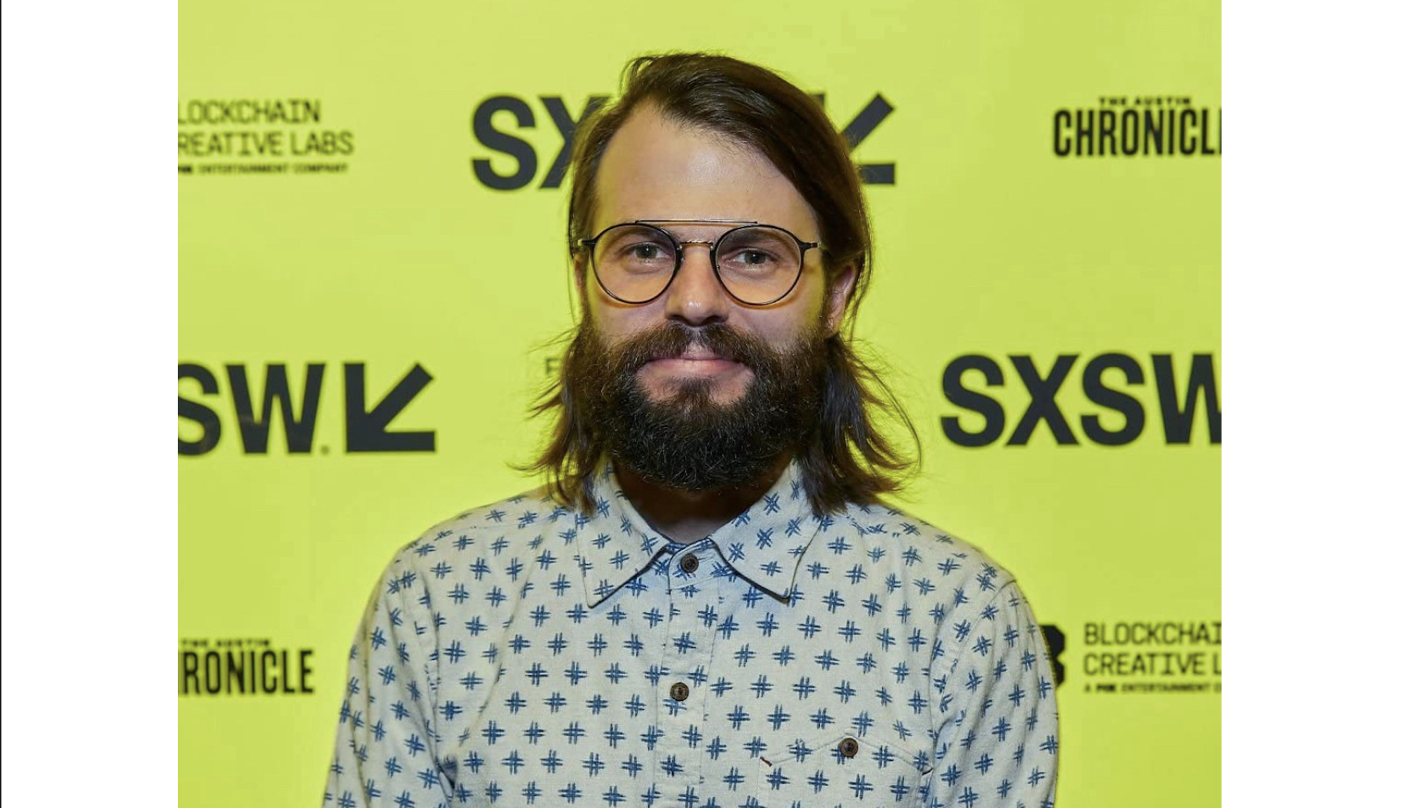 A color photo of Colin West, a white man with dark brown shoulder-length hair and facial hair, in front of a bright yellow background with the SXSW logo on it.