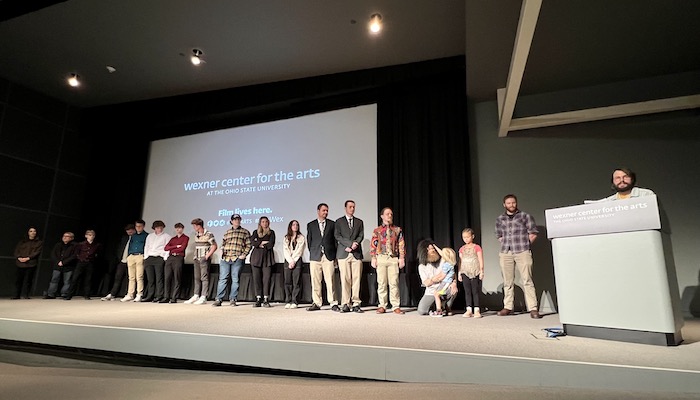 A lineup of 18 individuals on an auditorium stage with a large screen behind them. The screen holds a logo for the Wexner Center for the Arts. To the right of the lineup, another man stands on stage and speaks from behind a podium.