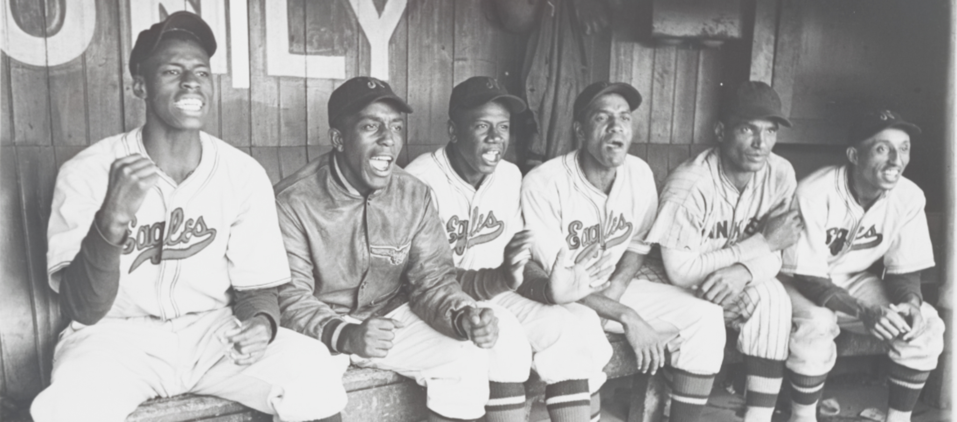 Six Black men in Newark Eagles baseball uniforms excitedly cheer on their team from a bench in the dugout.  