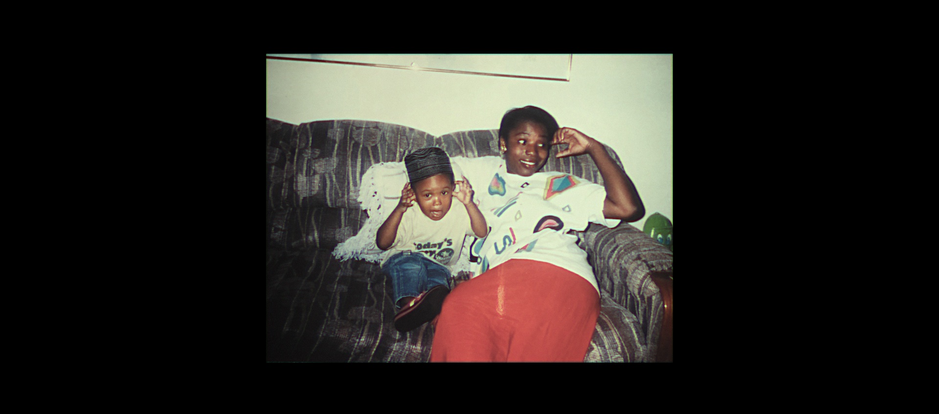 A vintage color photo of a young, smiling Black woman and her small son sitting on a couch. The image is surrounded by a thick black border.