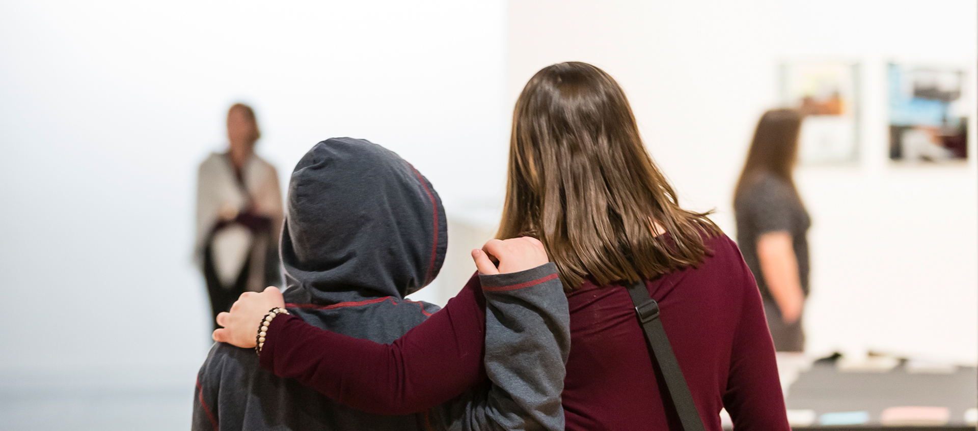 Two people viewed from behind stand in the galleries with their arms around each other. Two artworks hang on a white wall in the blurred background.