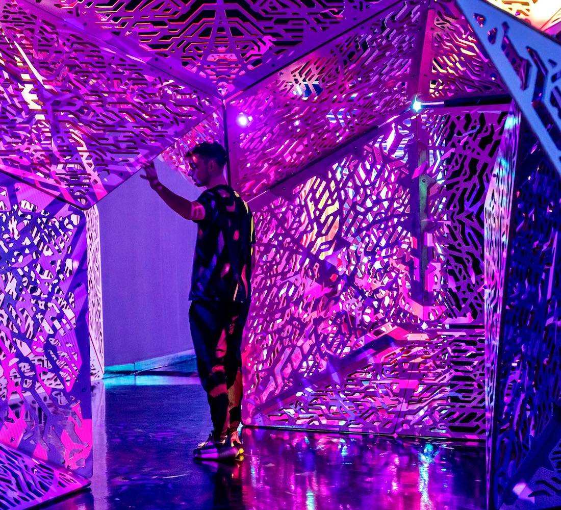A man walking through an immersive art space. He’s surrounded by abstract geometric forms lit in pink and blue