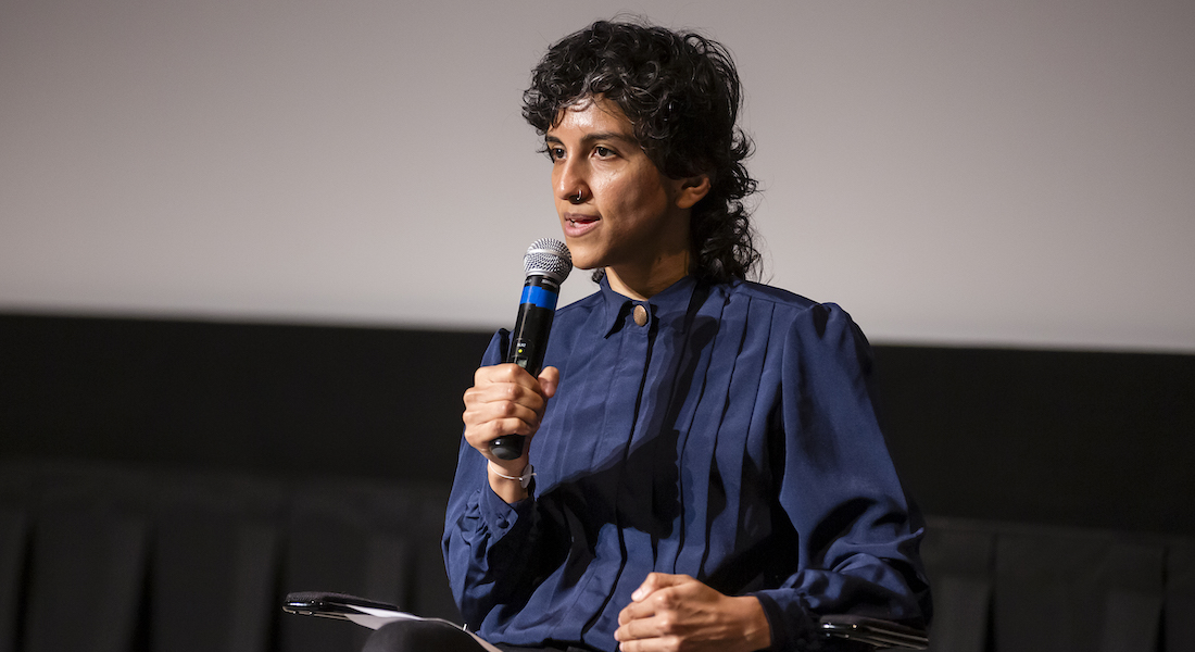 A non-binary person of Pakistani descent sits on a stage and speaks into a microphone