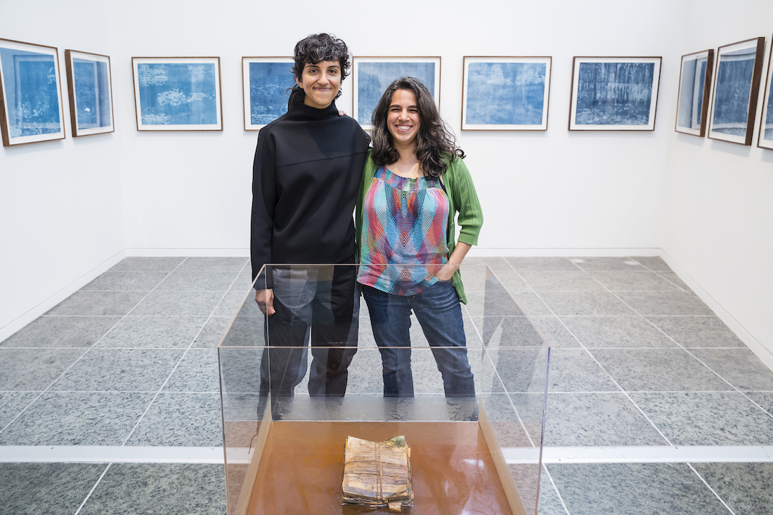 Two siblings stand together smiling in a gallery space with blue prints on the walls.
