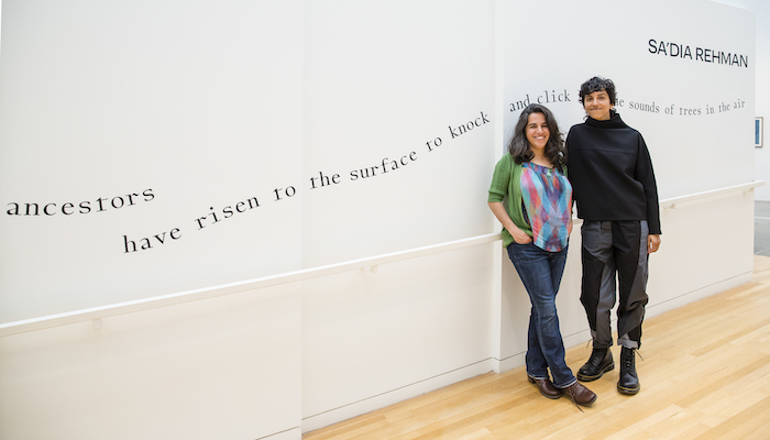 Two siblings Stand next to each other against a gallery wall, smiling.