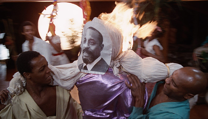 Two Black people hold up an effigy that is on fire, a photo of a Black man is attached to the face of the effigy.