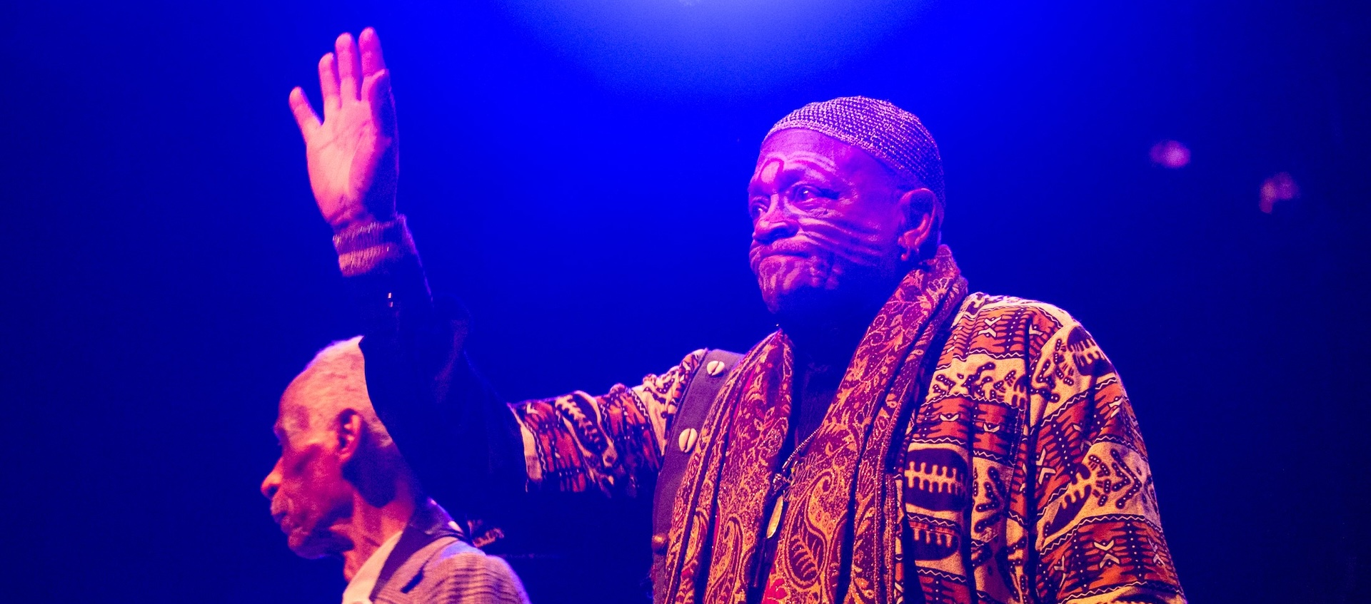 Two older men stand on stage under a blue light, one is wearing tribal face paint and waiving.  