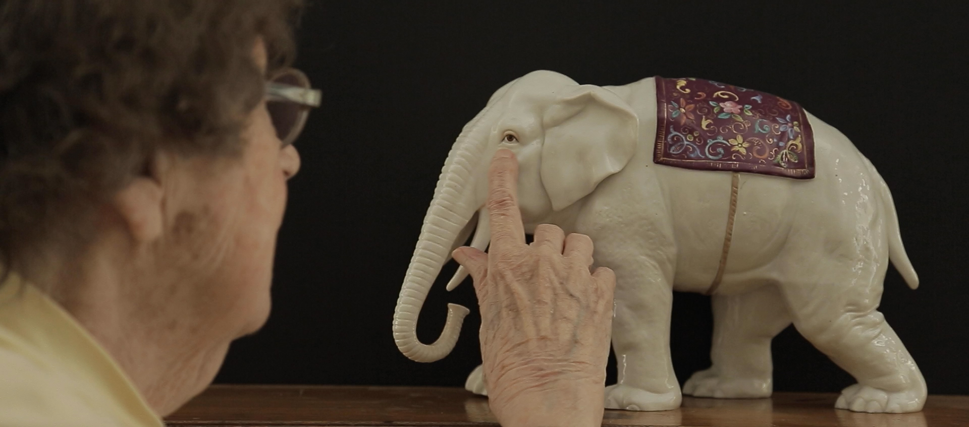 An older woman is touching the eye of a white elephant figurine.