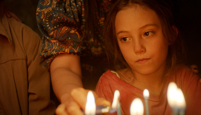 A child sits in front of a cake with candles, with an adult reaching beside them.