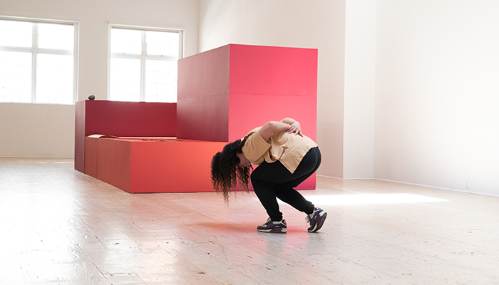 A long-haired dancer crouches in a gallery with their head toward the ground and arms behind them in front of three red platforms of differing heights.