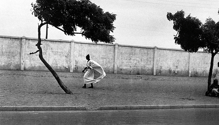 Panoramic photo of a Black person in a billowing white tunic walking next to a long, tall white wall. Two trees in the foreground frame the figure.