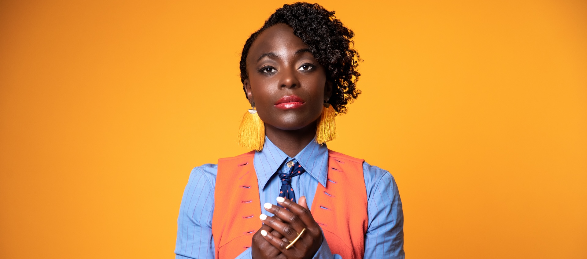 A woman with dark skin and short hair stands in front of an orange background with her hands clasped together as she stares at the camera. 
