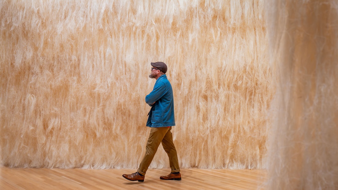 A man in a hat walks past a towering sculpture made of raw sisal.