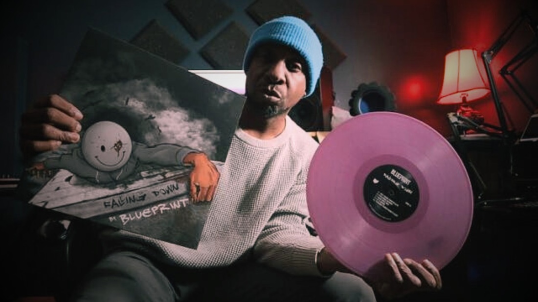 A man with dark skin and a knit beanie on his head holds out a pink vinyl album and accompanying cover.