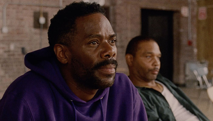 Two Black men sit in a large room, in the foreground wears a purple hoodie.