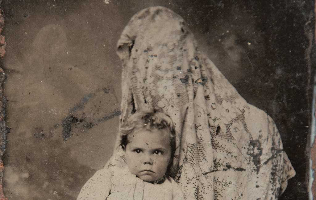 An antique black and white photograph of a small child looking solemnly at the camera. The child sits on the lap of someone completely covered in a floral wrap, who's there to hold the child still.