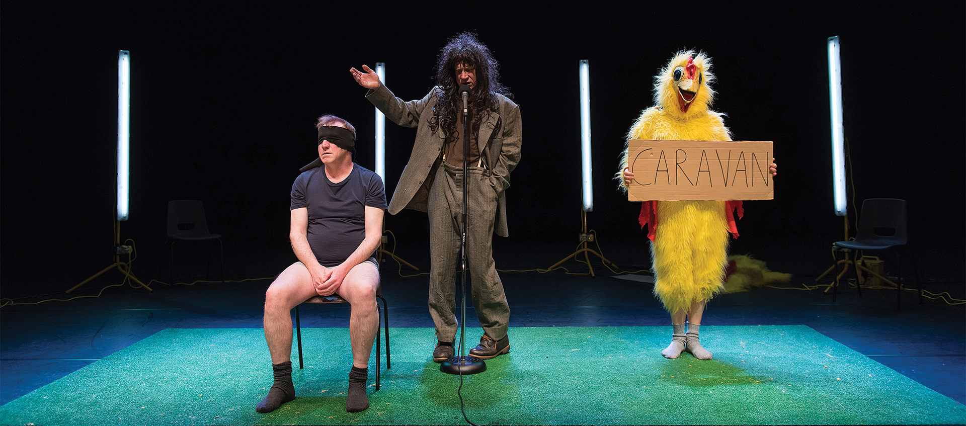 Performers on stage: A blindfolded man listens while another sings next to a performer in a chicken costume holding a sign that reads "caravan"