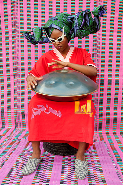 Video still of a black woman in a red robe and white sunglasses playing a steel drum against a vibrant pink background