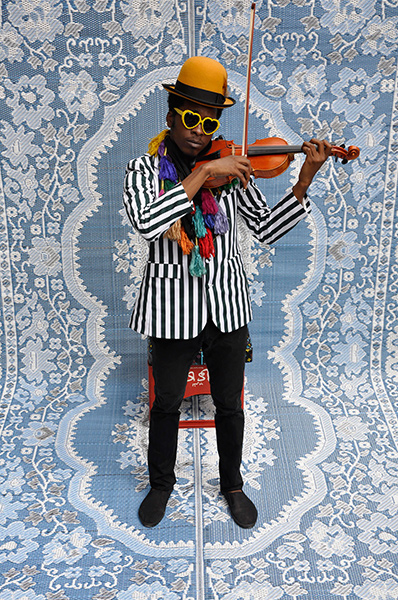 Video still of a musician in a a yellow hat and sunglasses playing the violin against an elaborate blue and white background