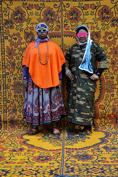 Video still of two women in headdresses and colorful sunglasses against an ornate background