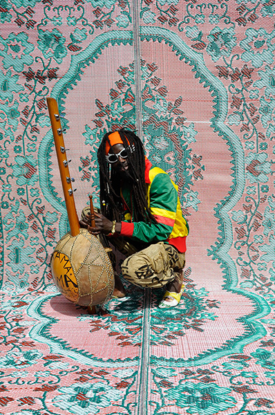 Video still of a dreadlocked man in green, yellow, and red clothing with a musical instrument