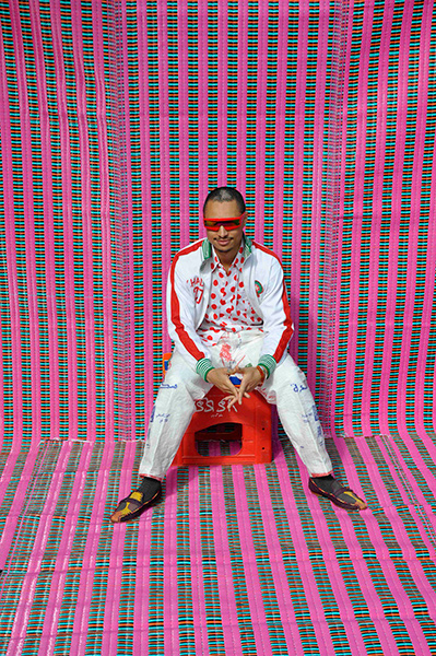Video still of a man in a white track jacket and red visor sunglasses against a bright pink background