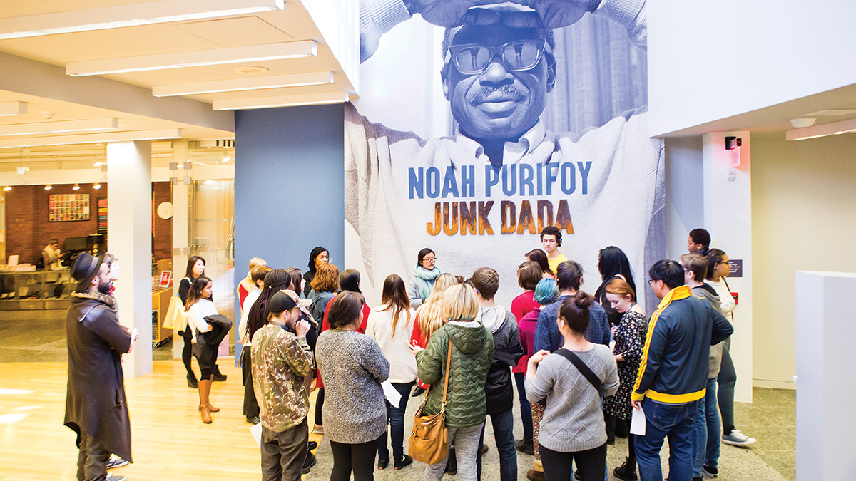 Title wall of the exhibition featuring a large mural of artist Noah Purifoy wearing sunglasses with arms stretched overhead