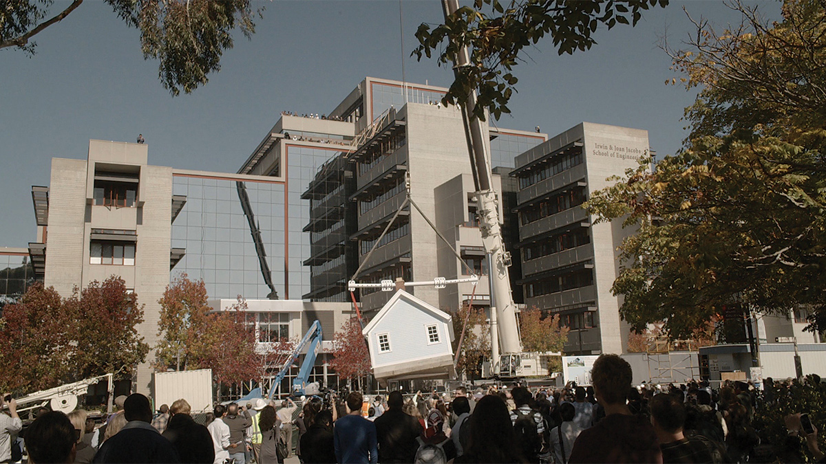 Film still of a crowd gathered to watch as a house is lifted into the air by a crane
