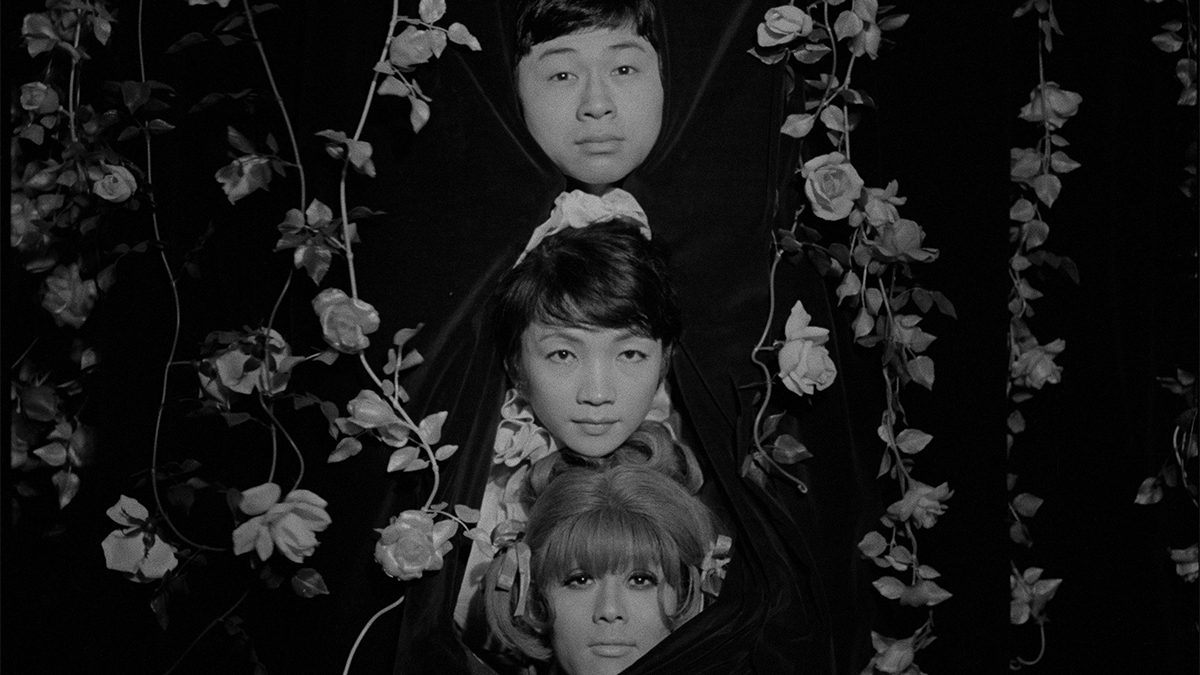 Film still of three people peaking out from a rose bush.