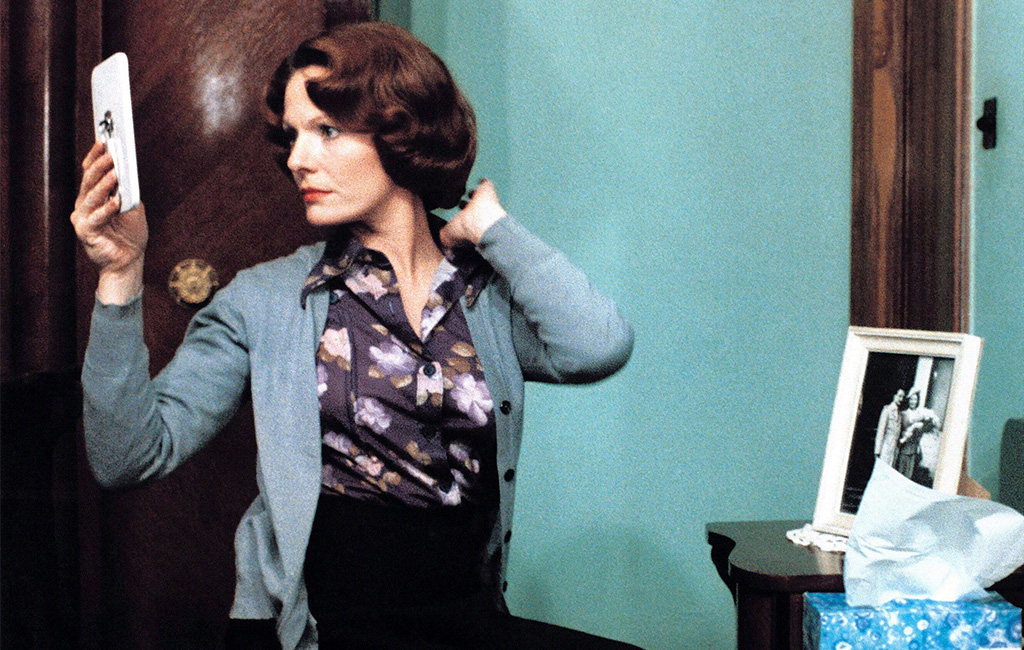 Still from the film Jeanne Dielman, 23 quai du Commerce, 1080 Bruxelles. A red headed woman sits at a table with a framed photograph sitting on it. She's wearing a patterned blouse and gray cardigan. She looks to the side, into a mirror, and fluffs her hair with her free hand.