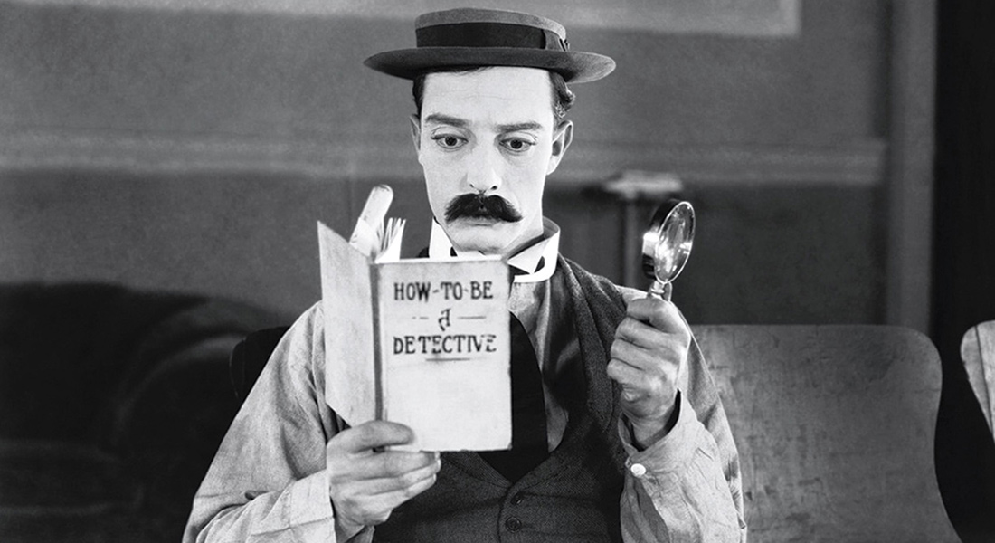 A man dressed like Sherlock Homes with a hat and mustache reads a book while holding a magnifying glass