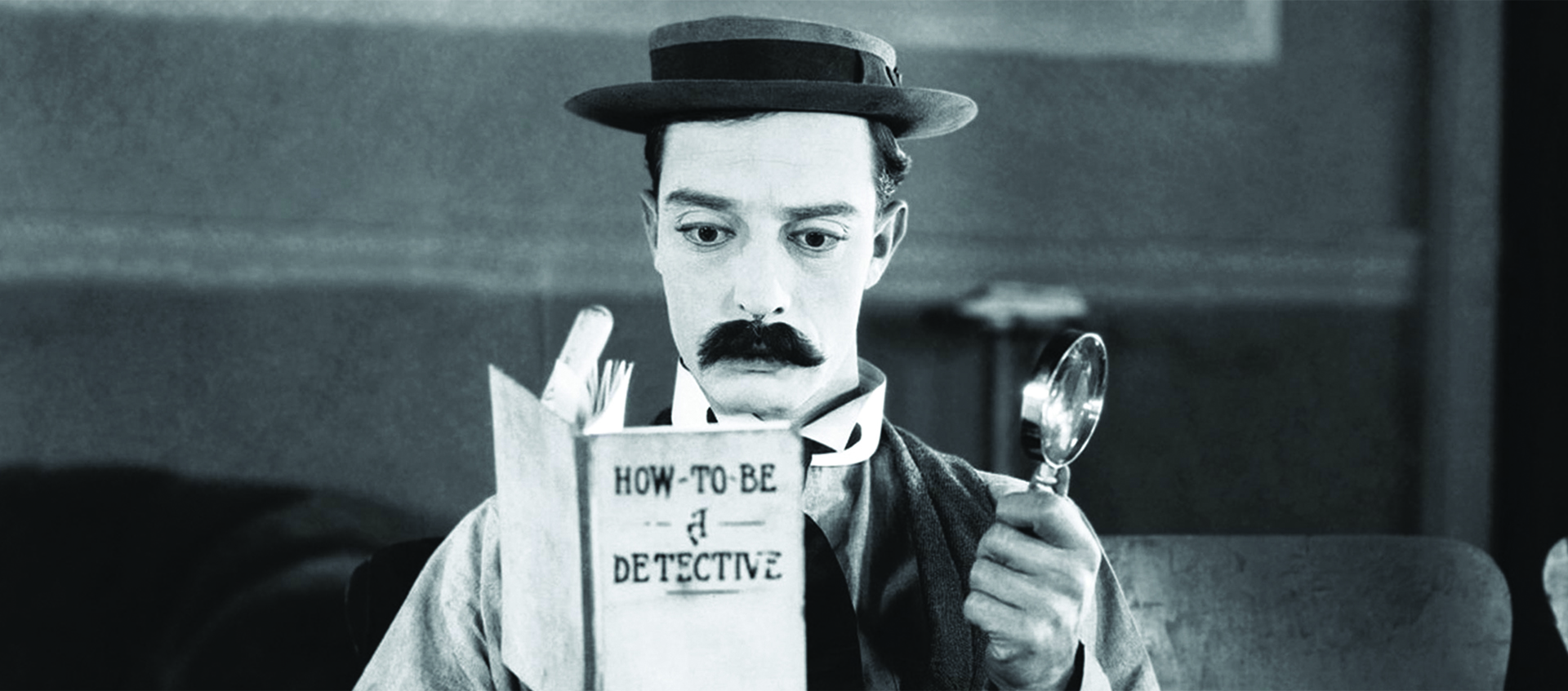 A man dressed like Sherlock Homes with a hat and mustache reads a book while holding a magnifying glass